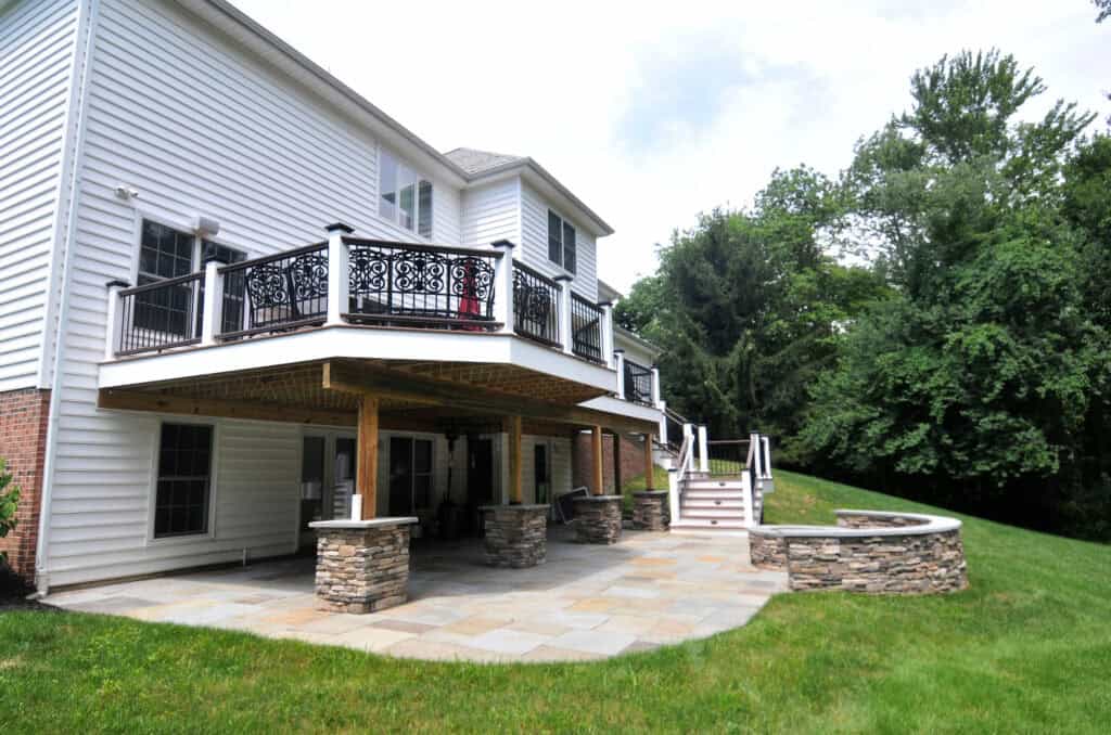 A two-story house with a white exterior features a second-floor balcony with ornate black railings, a covered stone patio below, and a built-in stone fire pit and seating area, overlooking a grassy yard and trees.