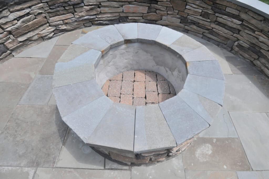 A circular stone fire pit with a brick-lined base, surrounded by a patio and a curved stone wall. The fire pit is empty and the surrounding area is clean.
