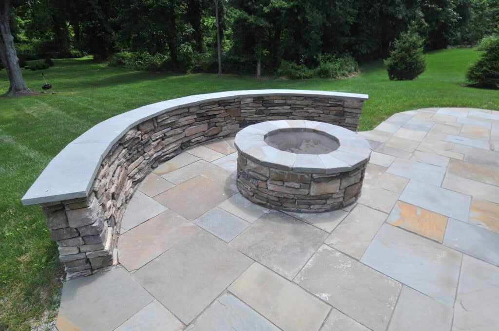 Curved stone bench and circular stone fire pit on a large stone patio, surrounded by green grass and dense trees in the background.