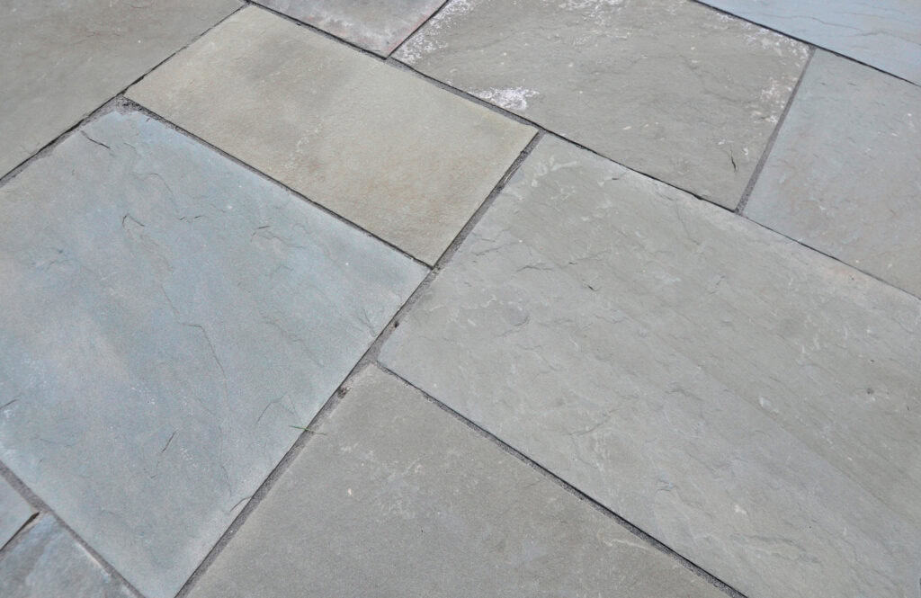 Close-up view of a stone tile floor with large, rectangular gray and beige tiles arranged in a staggered pattern. The surface shows natural texture and slight color variations.