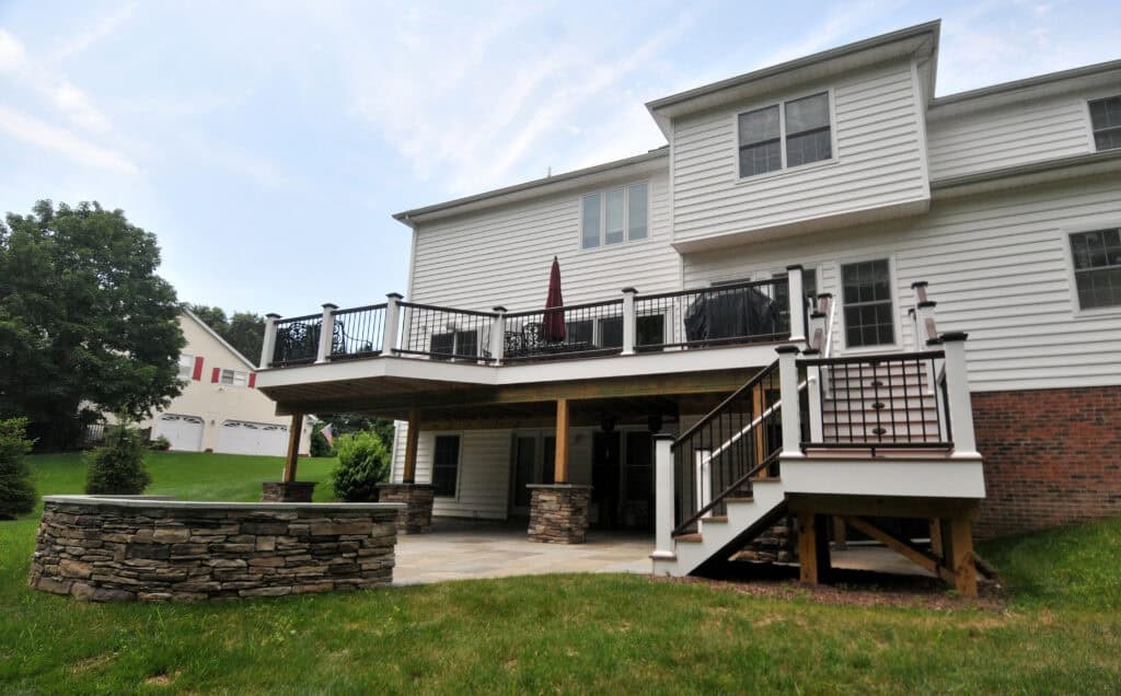 A two-story white house with a raised deck featuring black railings and an outdoor staircase. Under the deck is a covered patio area with stone columns. The yard is grassy and landscaped with trees and shrubs.