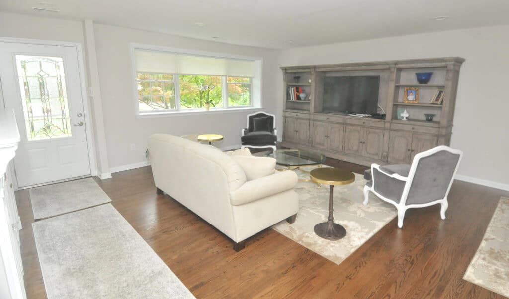 Bright living room with hardwood floors, a beige sofa, two gray armchairs, round side tables, a large wooden entertainment center with a TV, books, and decor, and a window letting in natural light.
