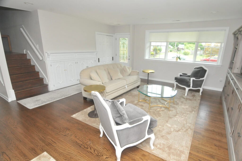A bright living room with hardwood floors, a beige sofa, two white armchairs with gray cushions, a glass coffee table, and large window letting in natural light. Stairs and a white door are visible in the background.