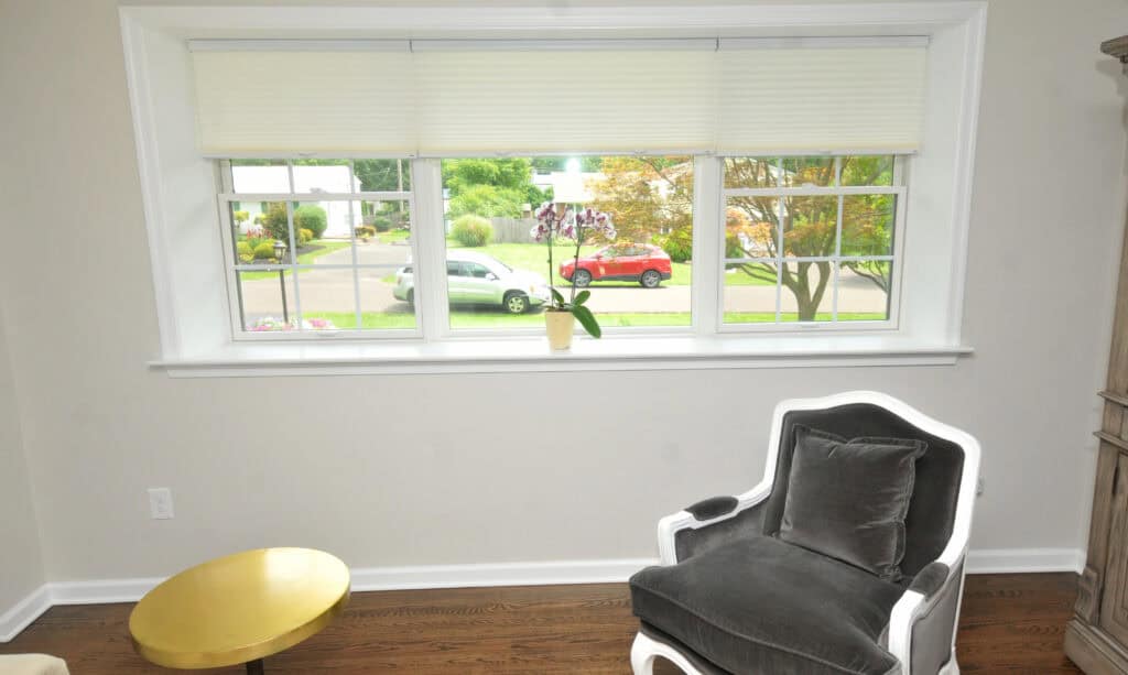 A bright room with a large window, gray armchair, small gold table, potted plant on the windowsill, and a view of parked cars and greenery outside.