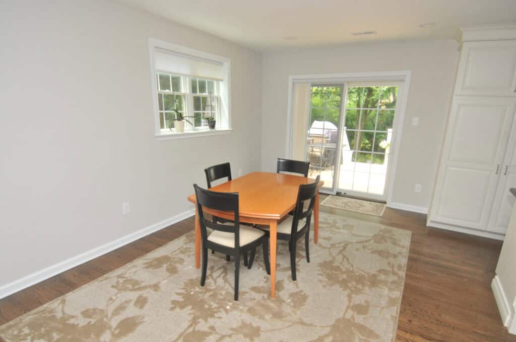 A dining area with a wooden table and four chairs on a patterned rug, next to a window and sliding glass doors that open to a patio with greenery outside.