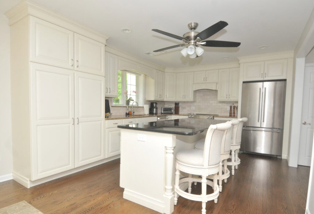 Bright, modern kitchen with white cabinets, stainless steel appliances, a central island with a dark countertop, and three upholstered barstools on wood flooring. A ceiling fan with lights hangs above the island.