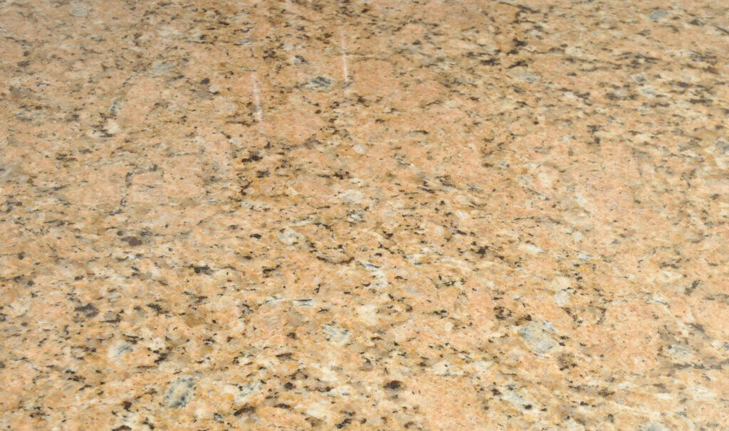 Close-up of a polished granite countertop with a beige base color and speckles of brown, black, and gray. The surface is shiny, reflecting some light.