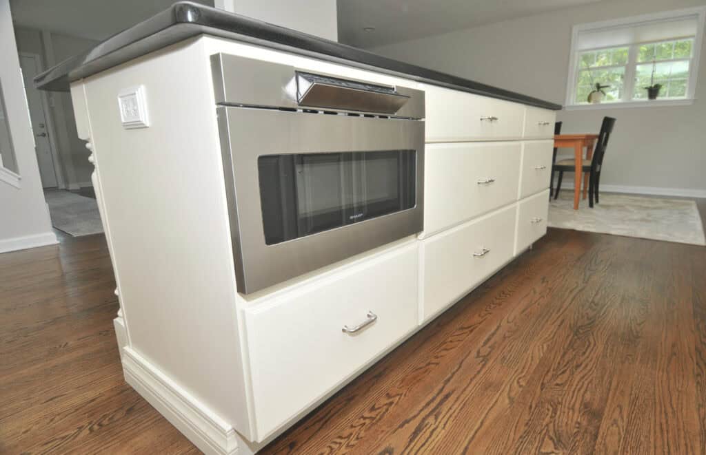 A kitchen island with white cabinets, a built-in stainless steel microwave, and a dark countertop, set on a hardwood floor. A dining table and window are visible in the background.