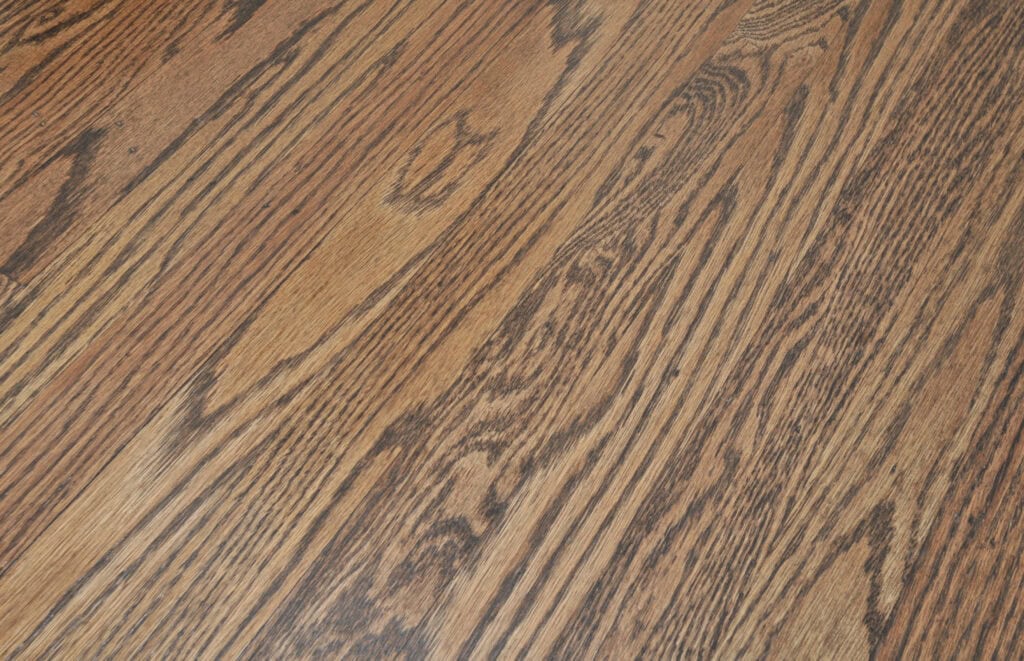 Close-up view of a wooden floor with a natural grain pattern and medium brown finish, showing the detailed texture and color variations in the wood planks.