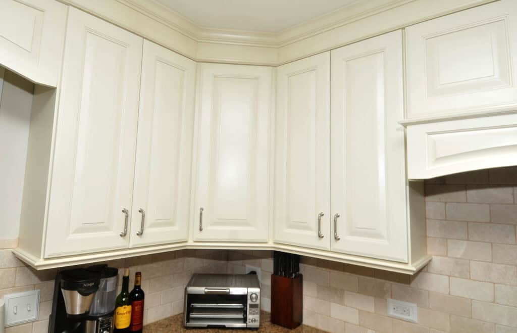 White kitchen cabinets with silver handles are mounted above a beige tiled backsplash. On the countertop below are a coffee maker, two bottles, a toaster oven, and a knife block with several knives.