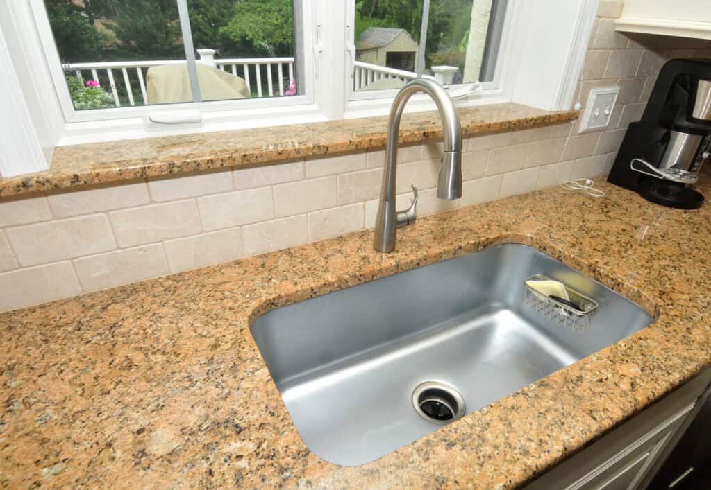 A clean stainless steel kitchen sink with a modern faucet set in a speckled granite countertop, beneath a window with a view of greenery outside. A sponge rests in a holder inside the sink.
