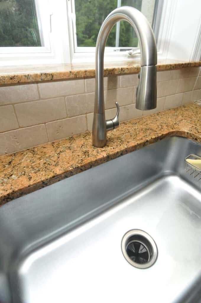 A stainless steel kitchen sink with a curved, brushed metal faucet on a granite countertop, set against a beige tiled backsplash and under a window with green trees visible outside.