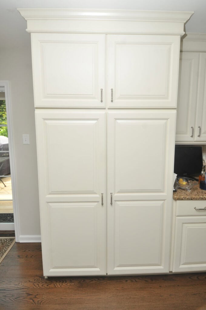 Tall, white kitchen pantry cabinet with four paneled doors and silver handles, standing on a dark wooden floor next to a granite countertop and smaller cabinets.