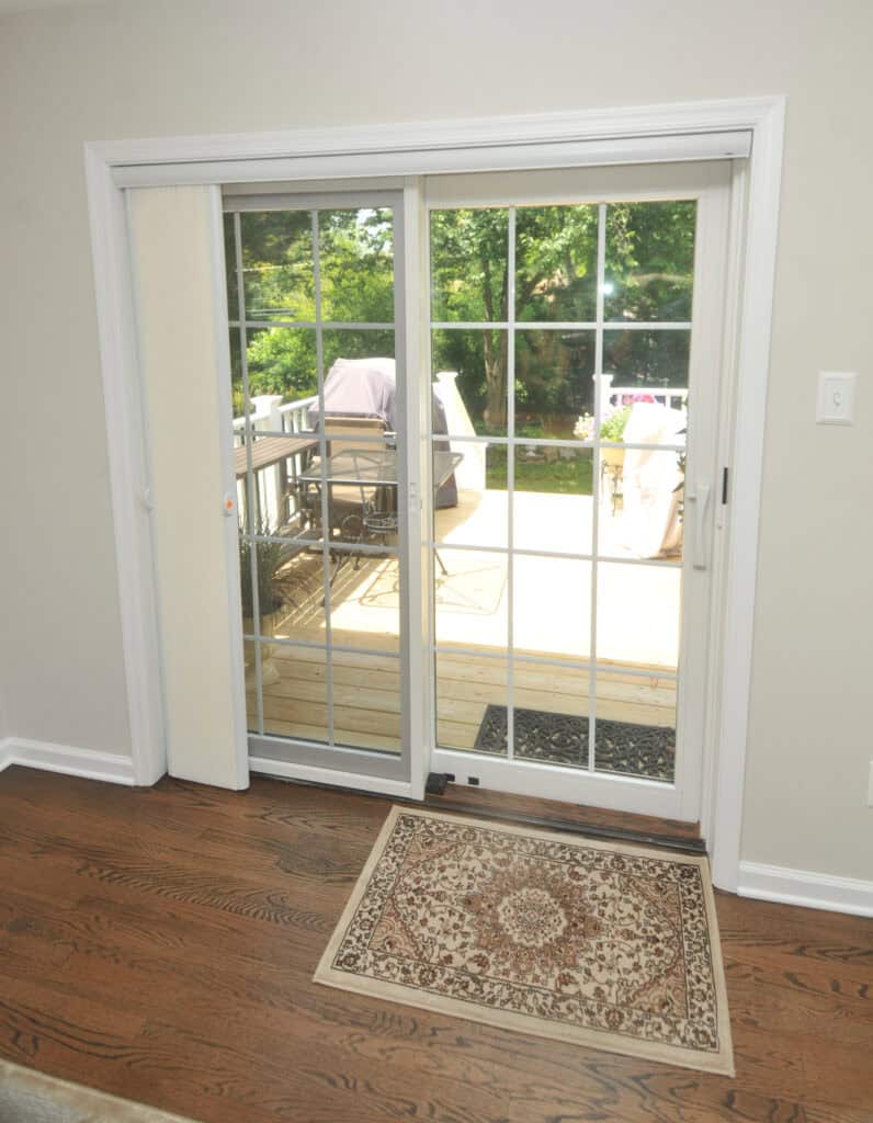 A glass sliding door with white grid frames opens to a wooden deck outside. A patterned rug lies on a hardwood floor inside, and outdoor furniture and greenery are visible through the door.