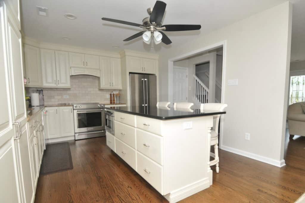 Modern kitchen with white cabinets, stainless steel appliances, a large island with drawers, two stools, hardwood floors, a ceiling fan, and neutral-colored walls. A staircase and living area are visible through open doorways.