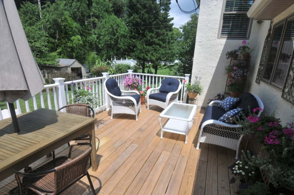 A wooden deck with white railings features cushioned wicker chairs, a glass-top coffee table, colorful potted plants, and a dining table with chairs. Lush green trees and a shed are visible in the background.