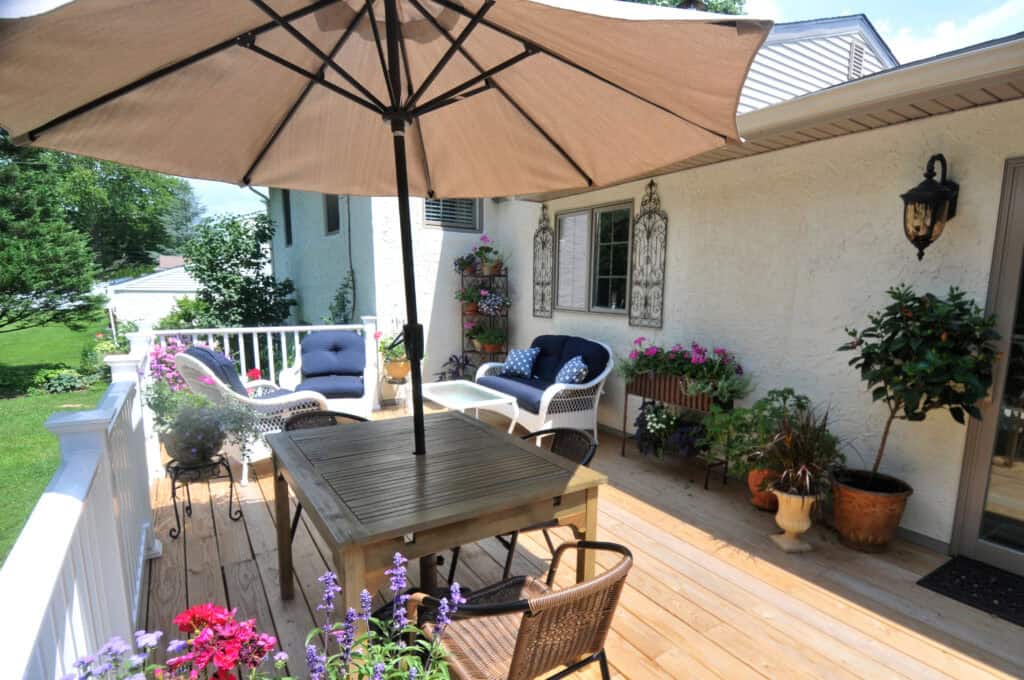 A cozy backyard deck features a wooden table with chairs under a large umbrella, cushioned seating, assorted potted plants, hanging baskets, and decorative wall art on a sunny day.