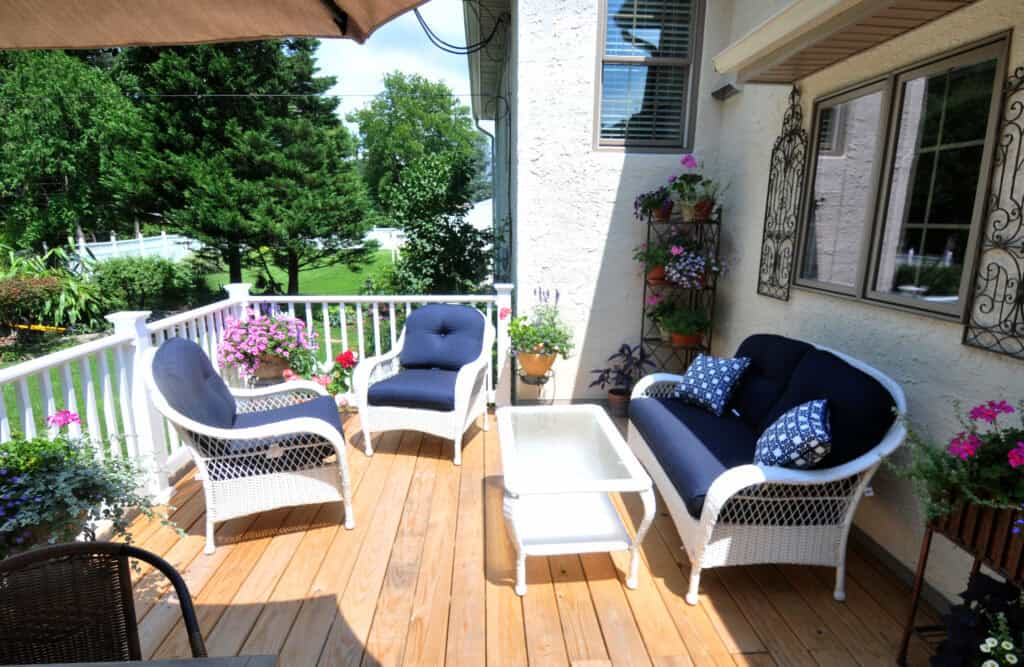 A cozy outdoor patio with white wicker furniture, blue cushions, a glass-top coffee table, potted plants, and flowers on a wooden deck, surrounded by a white railing and greenery.