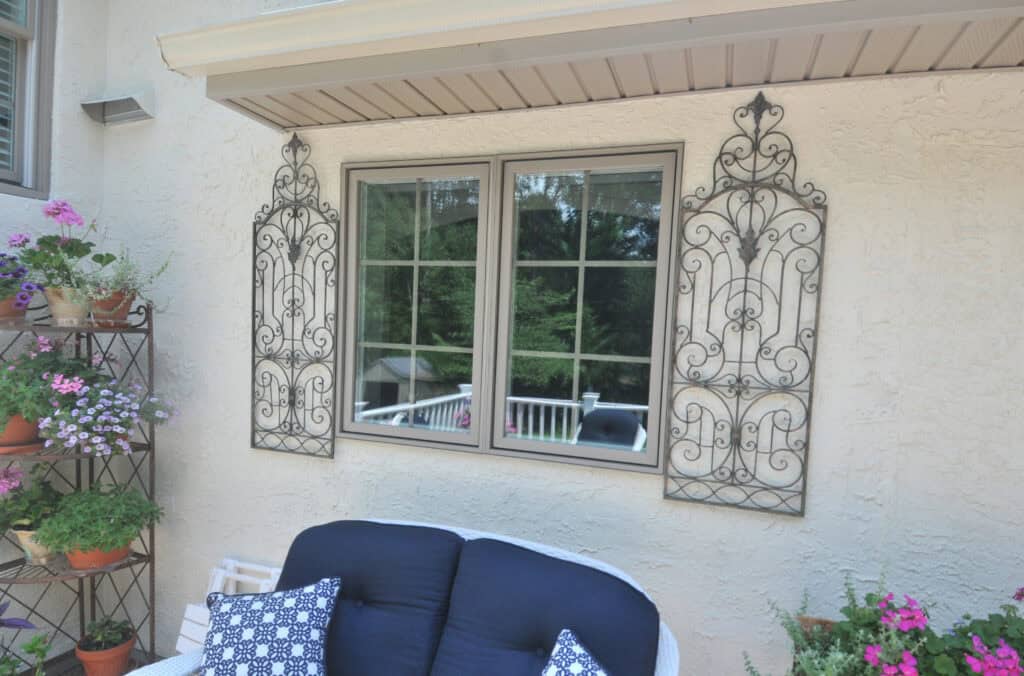 A cream-colored exterior wall with a double window, ornate black metal wall décor on each side, a black cushioned bench with patterned pillows, and a metal plant stand with potted flowers.