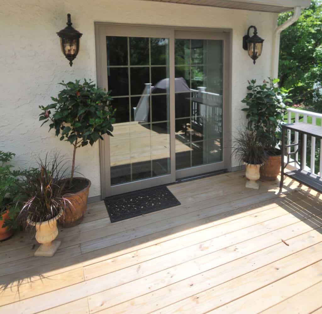 A wooden deck with potted plants on either side of sliding glass doors, two wall-mounted lanterns, a small doormat, and a grill and railing visible outside in the background.