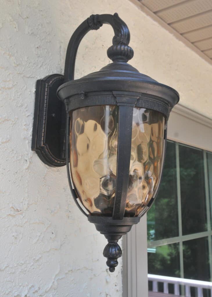 Black metal outdoor wall lantern with textured amber glass, mounted on a stucco wall next to a window reflecting trees and a railing.
