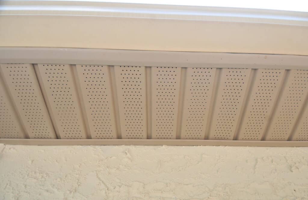 Close-up view of a beige perforated soffit vent under the eaves of a roof, with textured exterior wall visible at the bottom of the image.