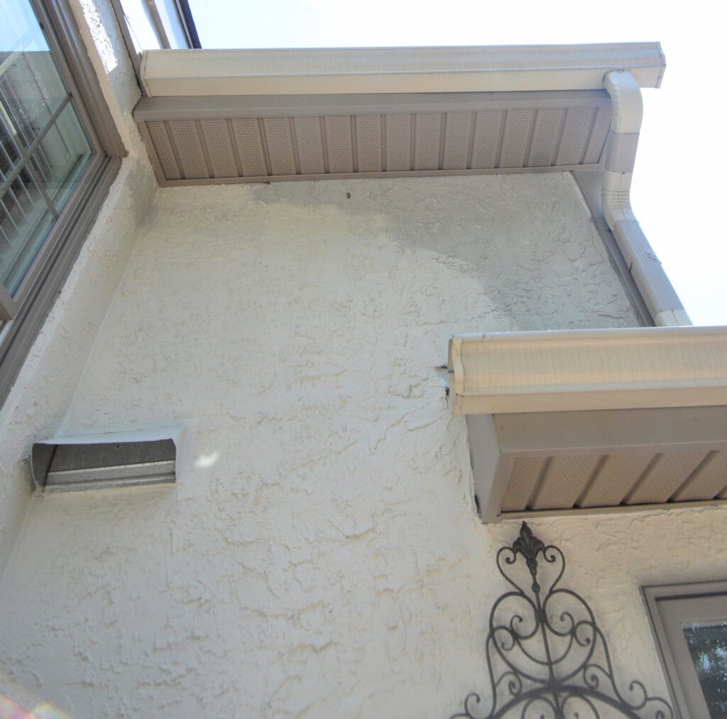 View looking up at the exterior corner of a stucco house with beige soffits, gutters, and a decorative metal wall ornament. A vent is visible on the left side of the wall near a window.