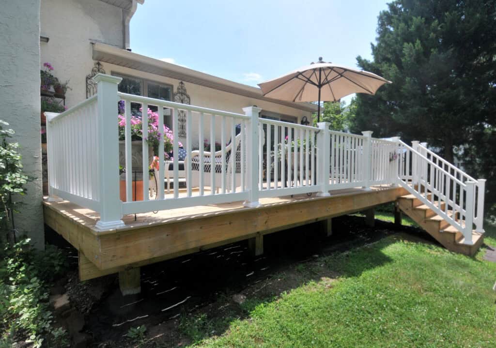 A raised wooden deck attached to a house, with white railings, stairs, potted flowers, outdoor furniture, and a large beige patio umbrella, overlooking a grassy yard with trees in the background.