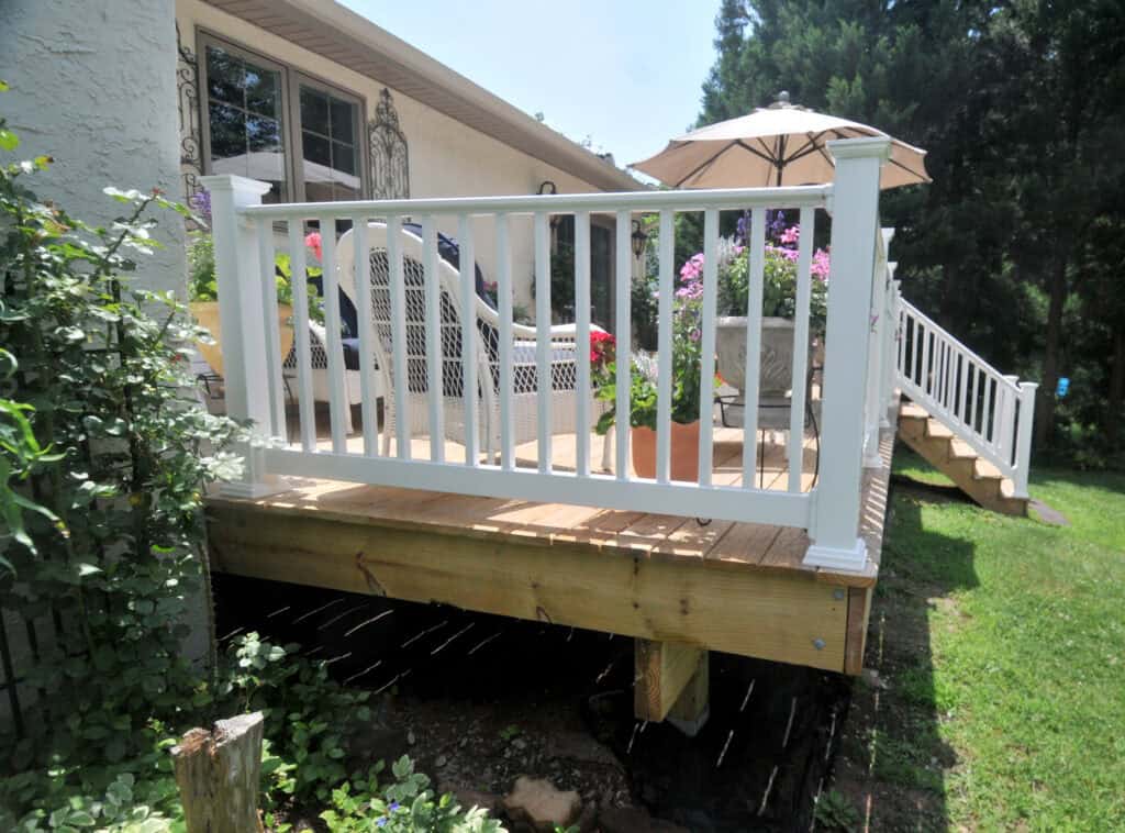 A backyard wooden deck with white railings, outdoor furniture, potted flowers, and an umbrella. The deck is elevated above grass and shaded by nearby trees. Steps lead down from the deck to the yard.