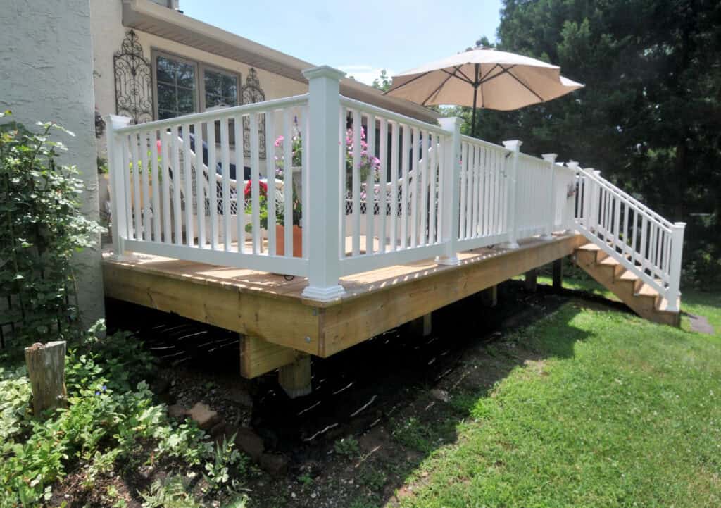 A wooden deck with white railing and stairs leads up to a house. Pink flowers in pots and a large patio umbrella decorate the deck. The surrounding area has green grass and leafy plants.