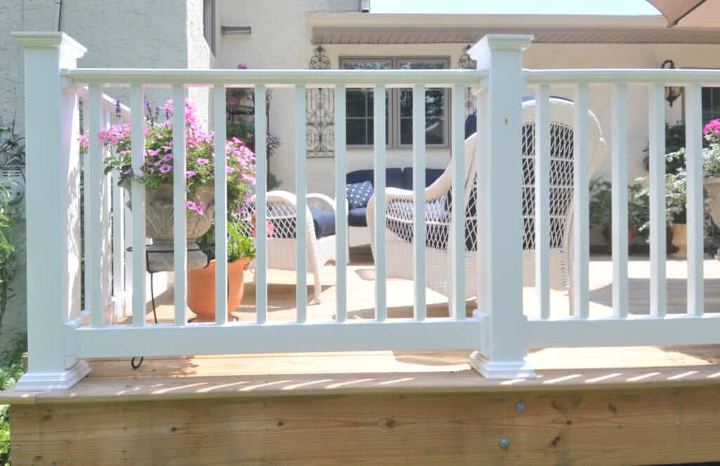 Sunlit patio with white railing, white wicker chairs, and potted pink flowers; wooden deck with a beige house in the background.