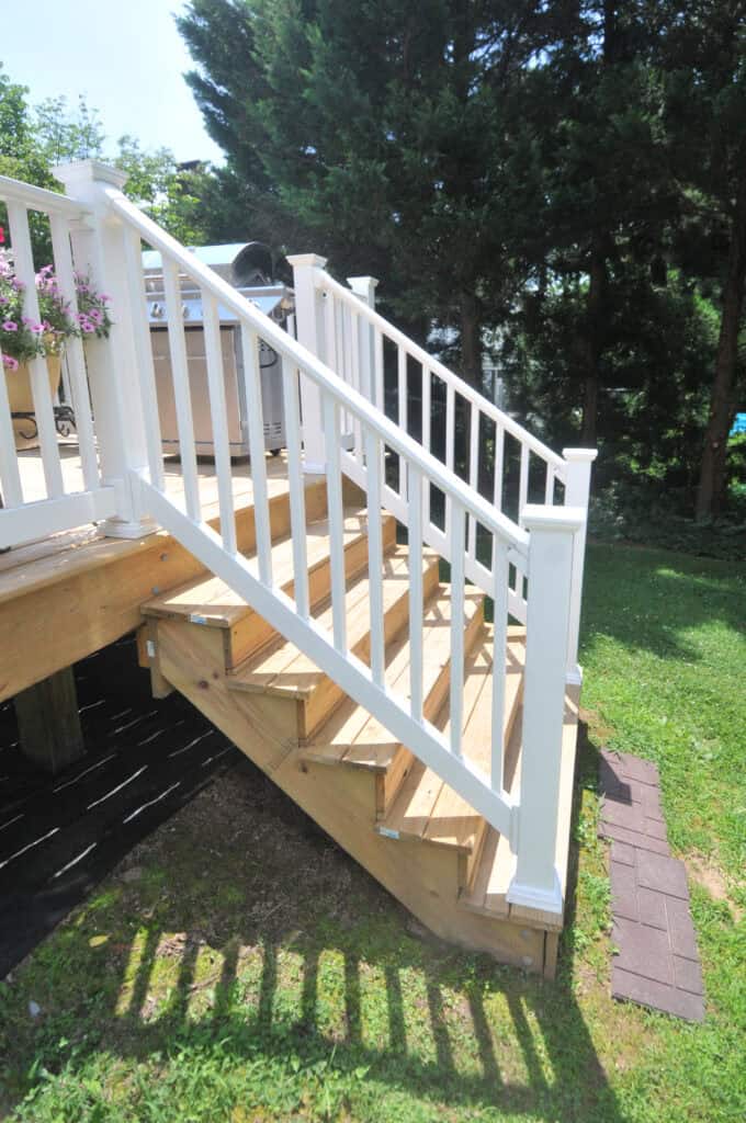 A wooden outdoor staircase with four steps and white railings leads from a deck to a grassy yard, surrounded by trees and greenery. Brick pavers form a small path at the bottom of the stairs.