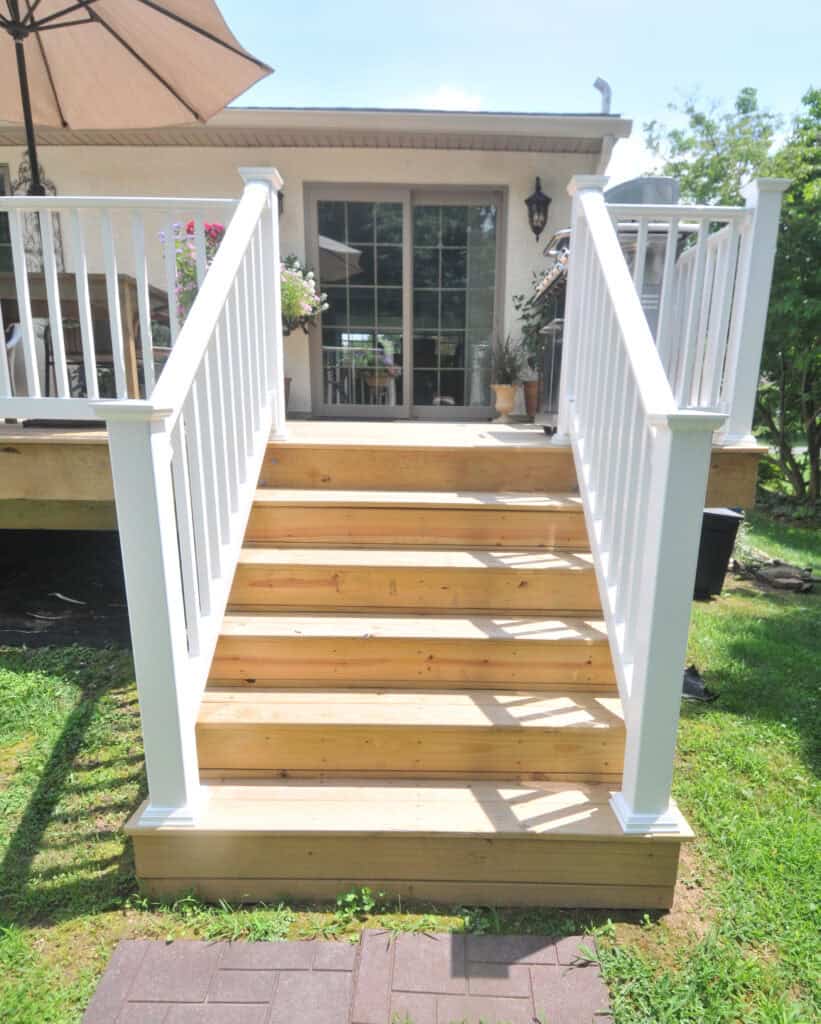 Wooden porch stairs with white railings lead up to a deck attached to a house. There is a glass door, outdoor furniture, an umbrella, and potted plants on the deck. Grass and a tree are visible in the yard.