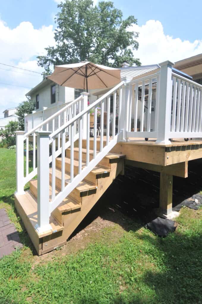 A wooden deck with white railings and stairs leads down to a grassy backyard. An umbrella is visible on the deck, and a house is in the background on a sunny day.