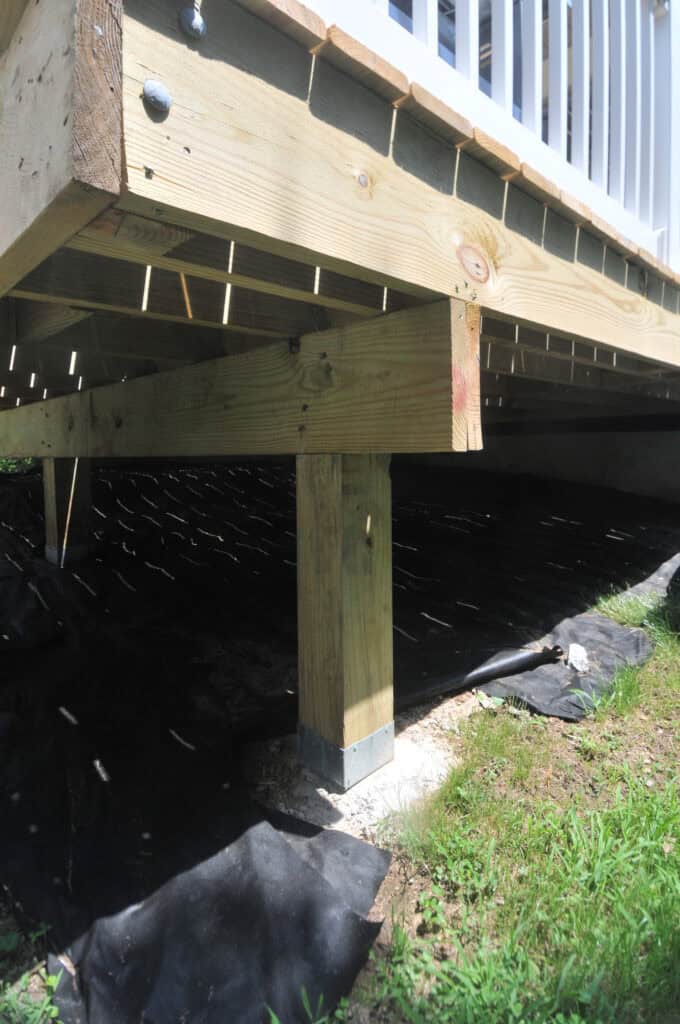 A close-up view of a wooden deck’s underside showing support beams and a vertical post anchored in a concrete base, with black tarp covering the ground beneath. Sunlight filters through the deck, creating dappled light spots.