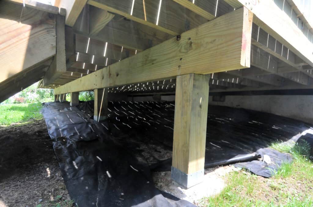 View under a wooden deck showing support beams, vertical posts on concrete footings, and black landscaping fabric covering the ground to prevent weed growth. Sunlight filters through the deck boards above.