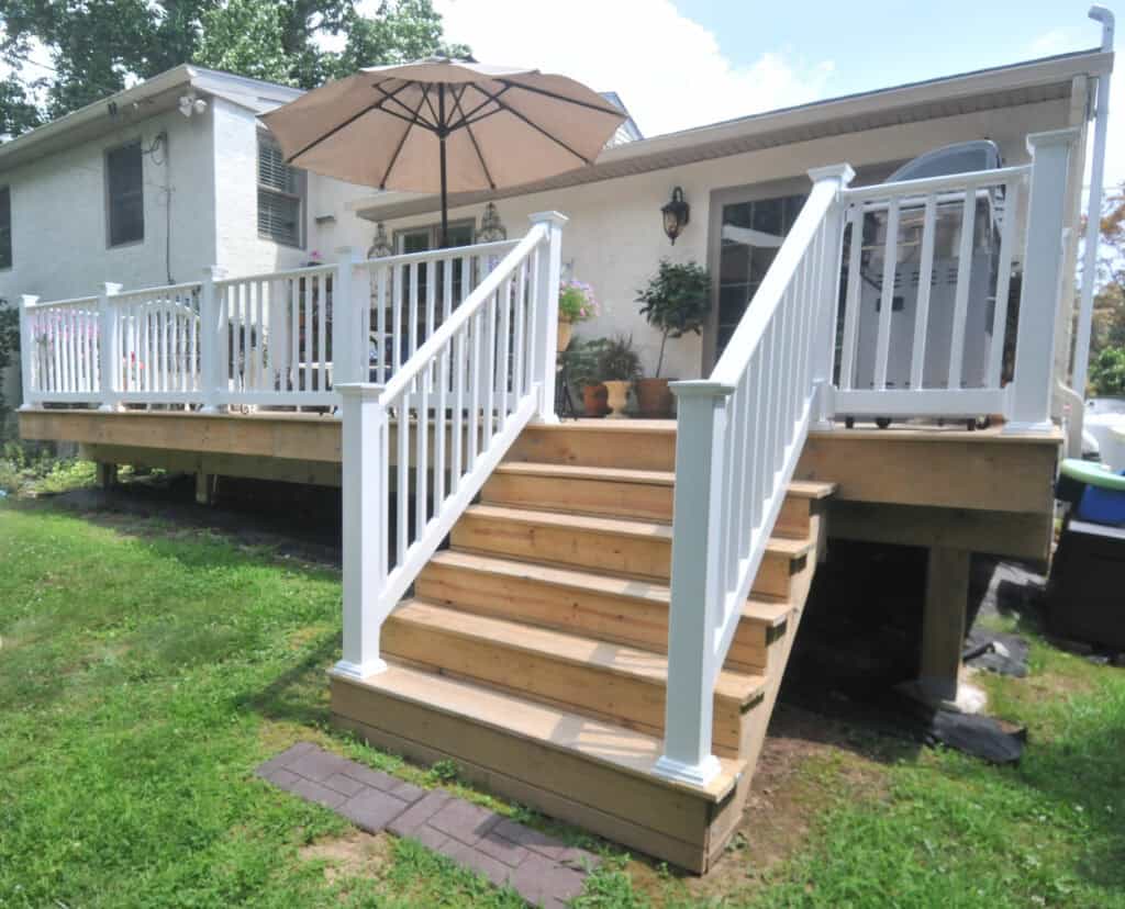 A wooden deck with white railings and stairs leads up to the back entrance of a house. There are potted plants, an umbrella, and an outdoor grill on the deck. The deck overlooks a grassy yard.