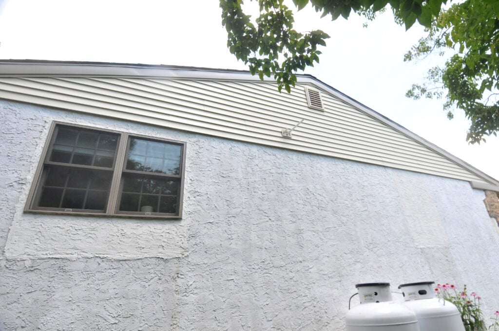 The image shows the side of a white stucco house with horizontal beige siding at the top, a two-pane window, two large propane tanks, and tree branches partially visible at the top.