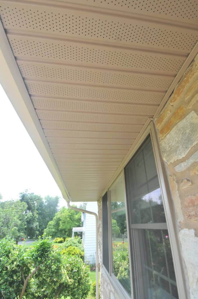 Close-up view of a beige soffit with vent holes under the roof eave of a house with stone siding and a window, overlooking a green yard with bushes and trees.