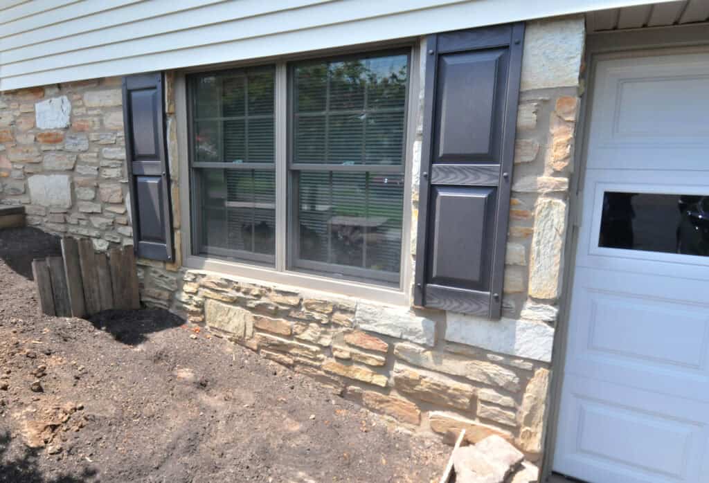 A stone house exterior with a large window featuring dark shutters, next to a white garage door. The ground in front appears freshly tilled or prepared for landscaping.
