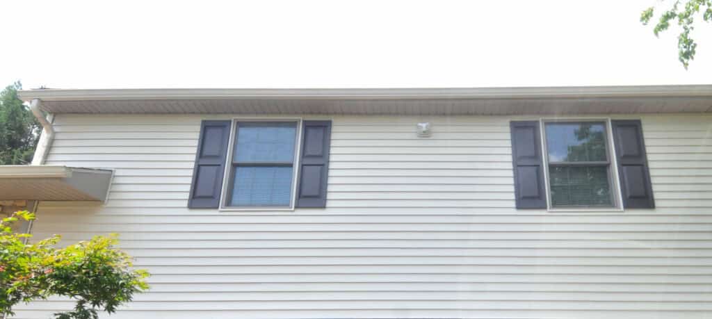 The upper exterior of a beige house with horizontal siding, two windows with dark shutters, and a small security camera or light fixture centered between the windows. Some tree branches are visible on the left and right edges.