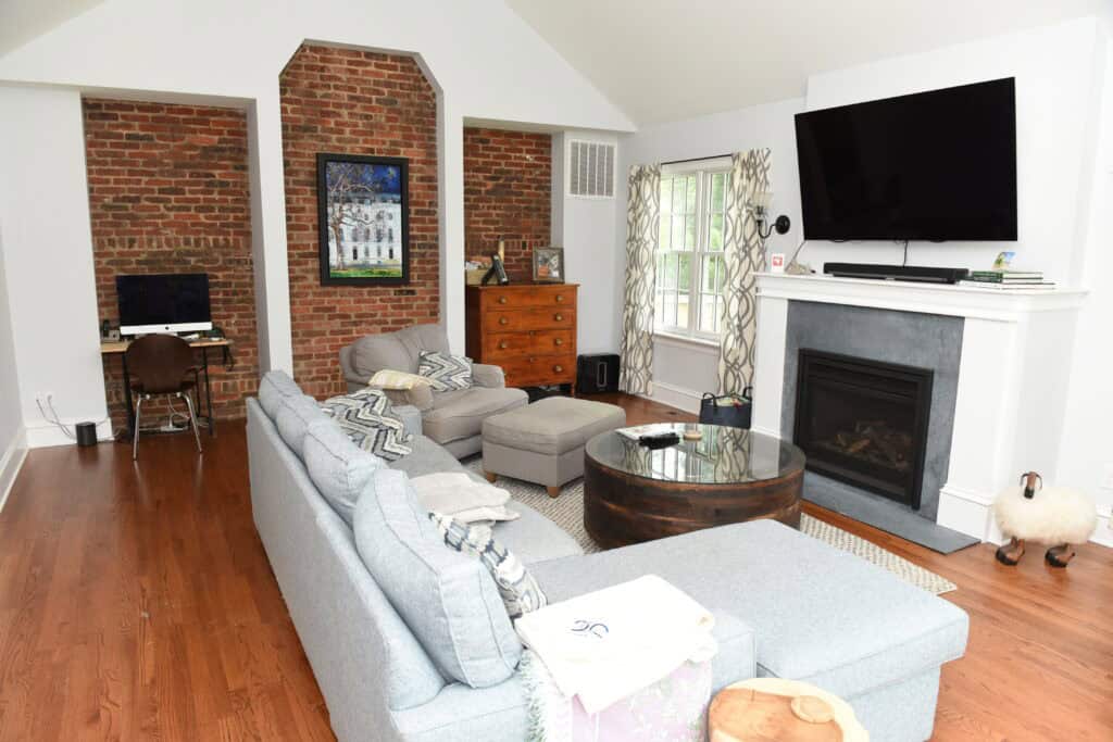A cozy living room with a gray sectional sofa, round coffee table, fireplace with a mounted TV, brick accent wall, small home office nook, dresser, and large window with patterned curtains.