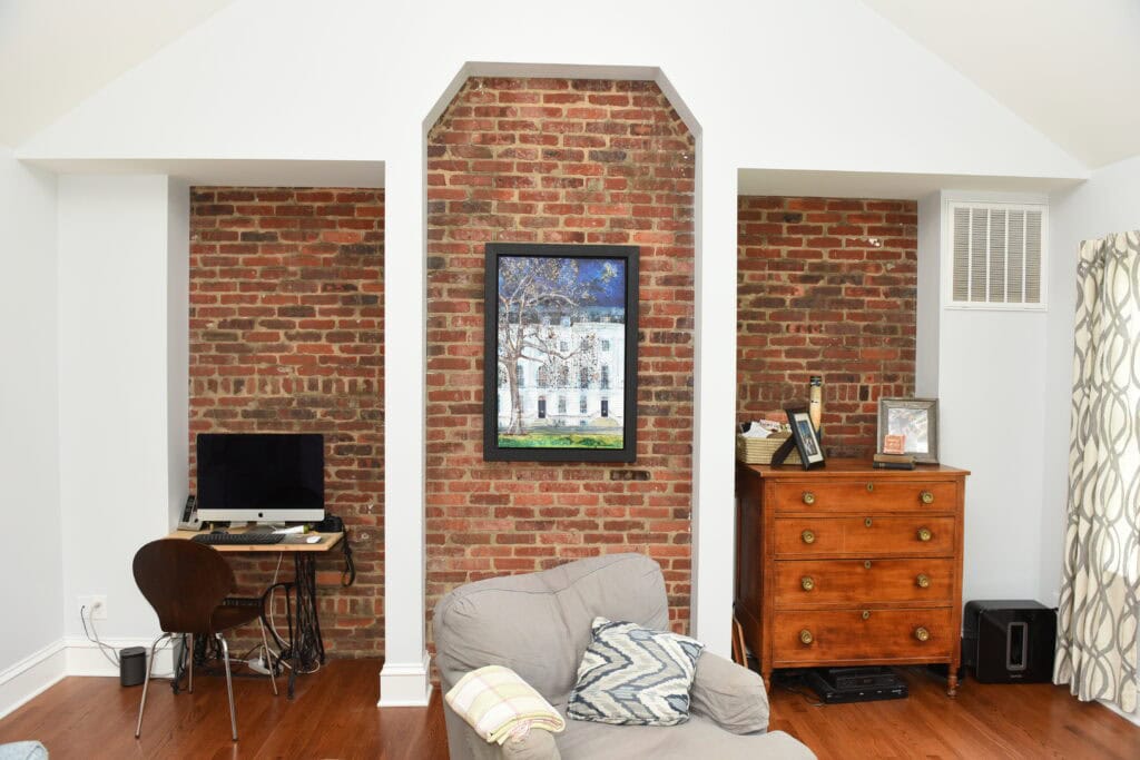 A home office and sitting area with exposed brick walls, a framed painting in the center, a computer desk on the left, a wooden dresser with decor on the right, and a gray armchair with pillows in the foreground.