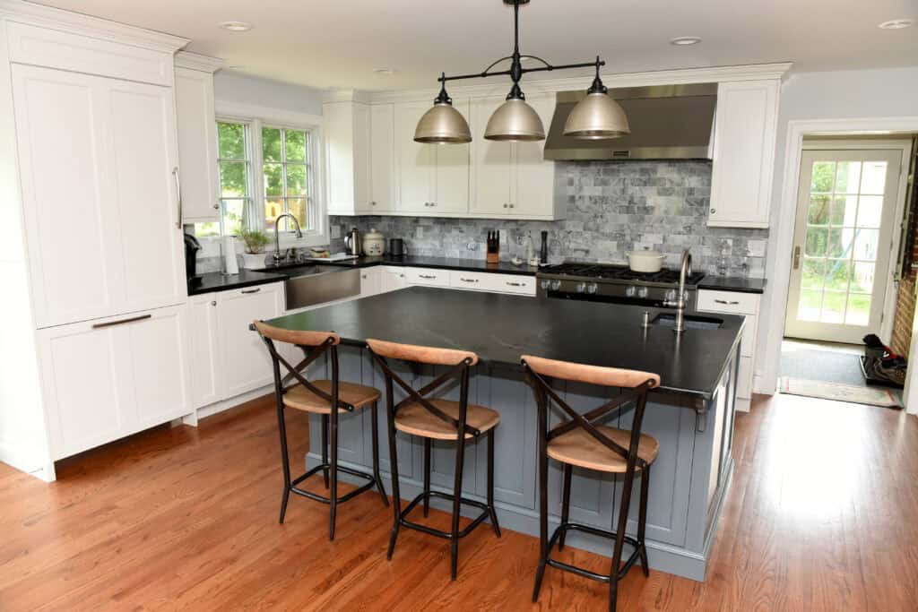 Modern kitchen with white cabinets, a large gray island with a black countertop, three wooden barstools, stainless steel appliances, a black backsplash, pendant lights, and hardwood floors. Sunlight streams in through windows.
