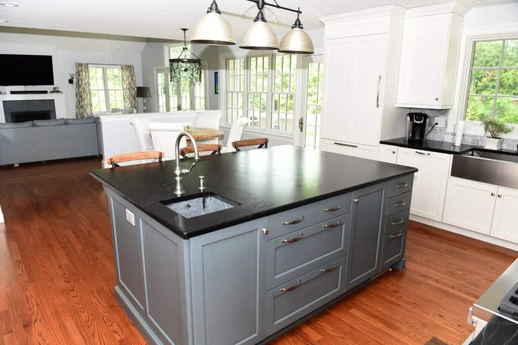 Modern kitchen with a large dark gray island, black countertop, sink, and barstools. White cabinets, hardwood floors, and bright natural light from large windows. Living and dining areas are visible in the background.