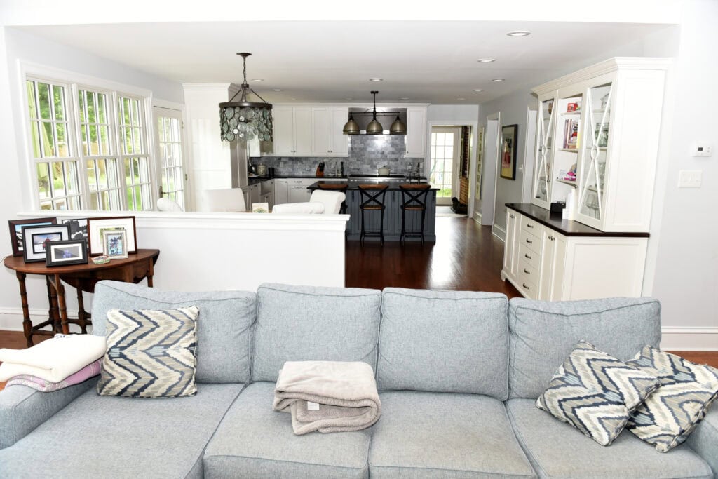 A bright, open living room with a light gray sofa, patterned cushions, and a folded blanket. Behind the sofa is a kitchen with white cabinets, a dark island, stools, and pendant lights. Sunlight streams through large windows.