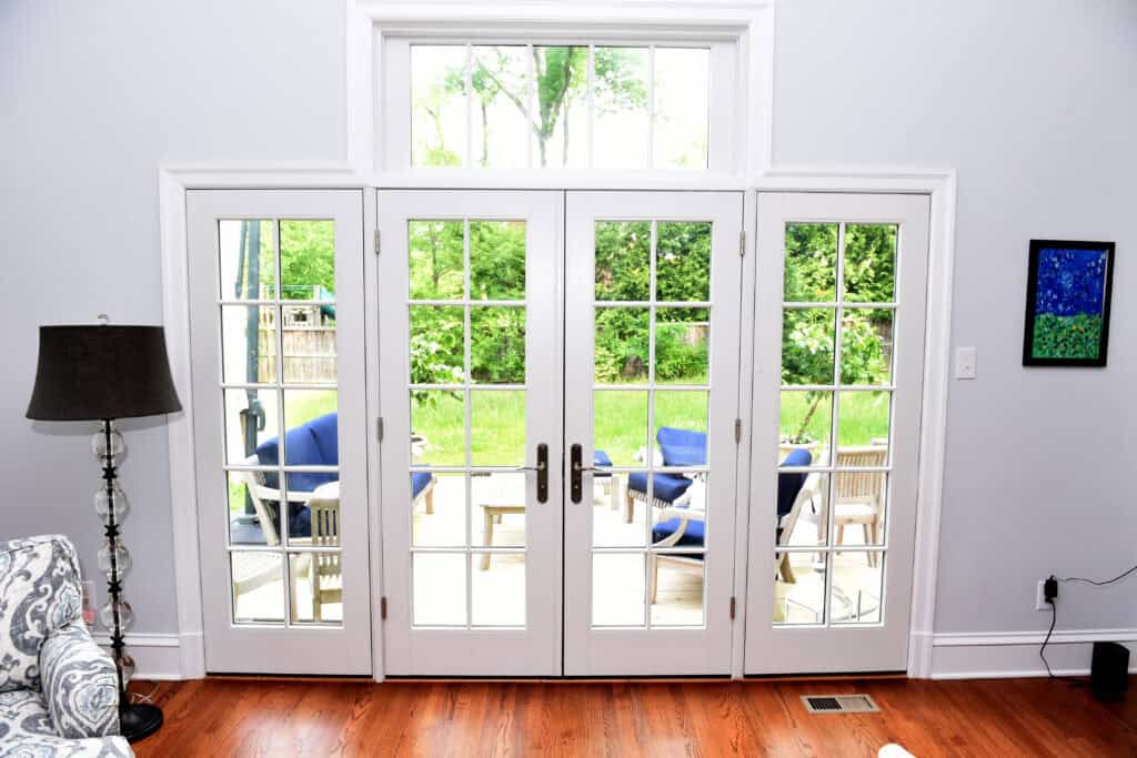 French doors with glass panes open to a patio furnished with blue cushioned chairs and a table. The room inside has wooden floors, a patterned sofa, a floor lamp, and a small colorful painting on a light gray wall.