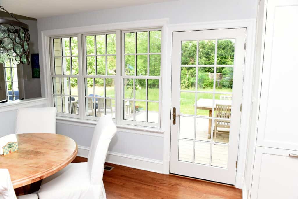 A bright dining area with a wooden table and white covered chairs near large windows and a glass door, looking out onto a green backyard with a patio table.
