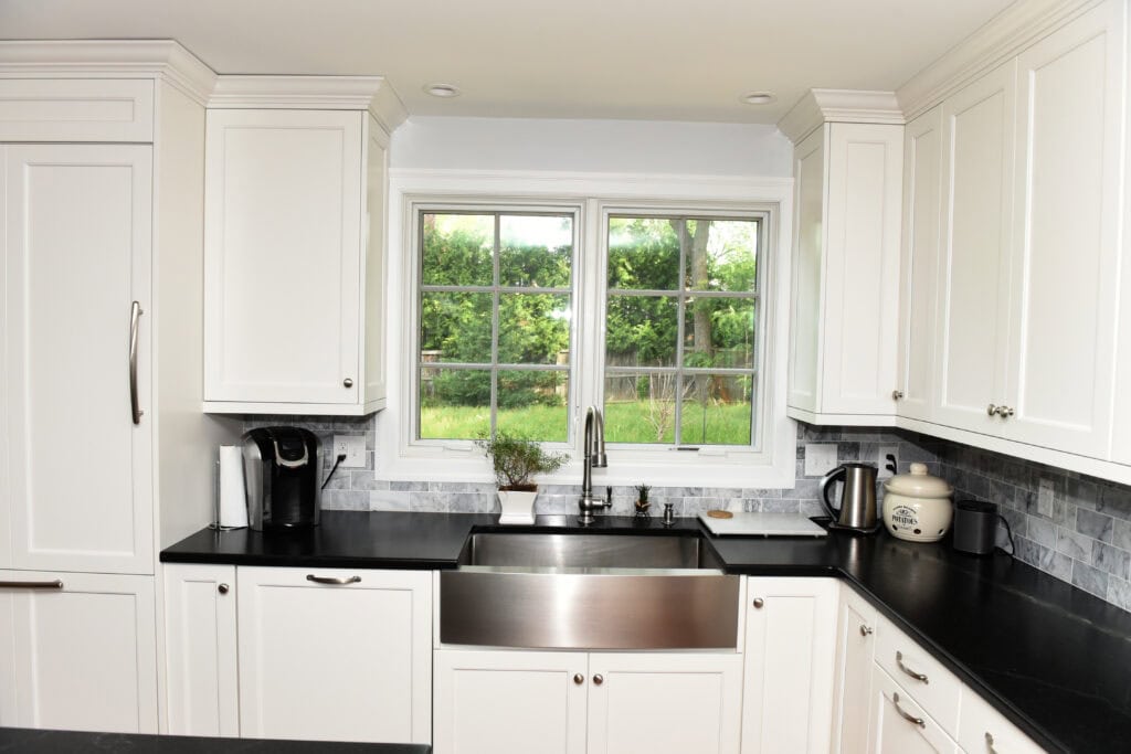 A modern kitchen with white cabinets, a large stainless steel farmhouse sink, black countertops, a coffee maker, kettle, and a window overlooking a green backyard.