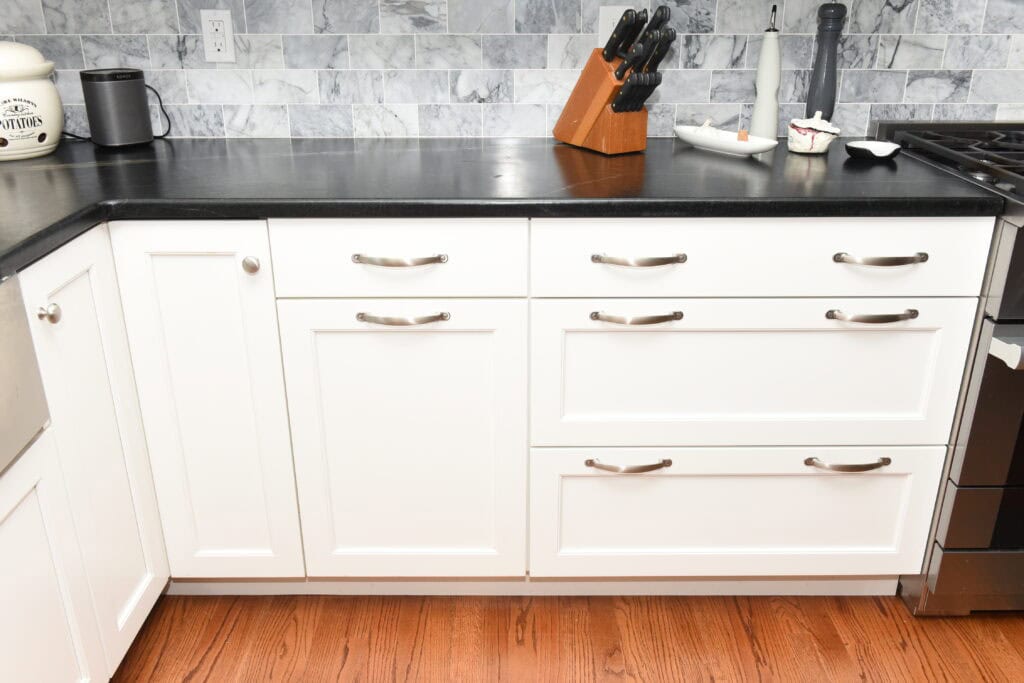 A modern kitchen with white cabinets, silver handles, black countertop, knife block, utensil holder, and a gray marble-tile backsplash. The floor is wood, and part of a gas stove is visible on the right.